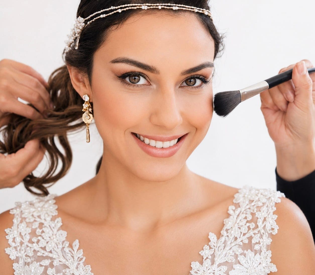 Novia sonriente con tocado de perlas mientras dos profesionales terminan su maquillaje y peinado
