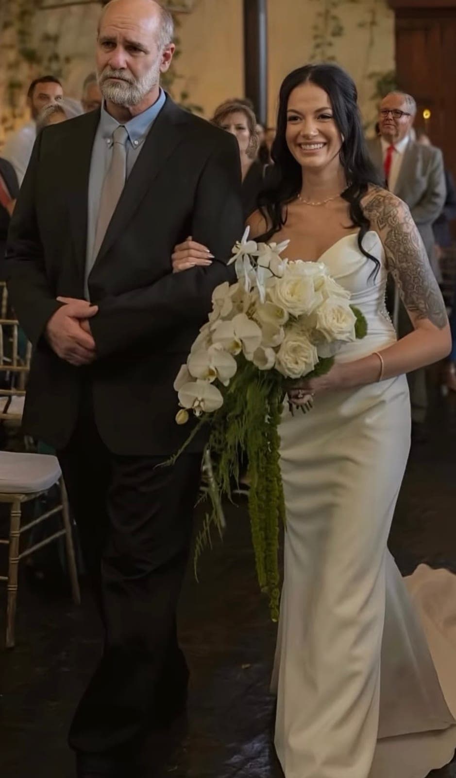 Novia entrando a la ceremonia del brazo de su acompañante, con ramo de novia en cascada