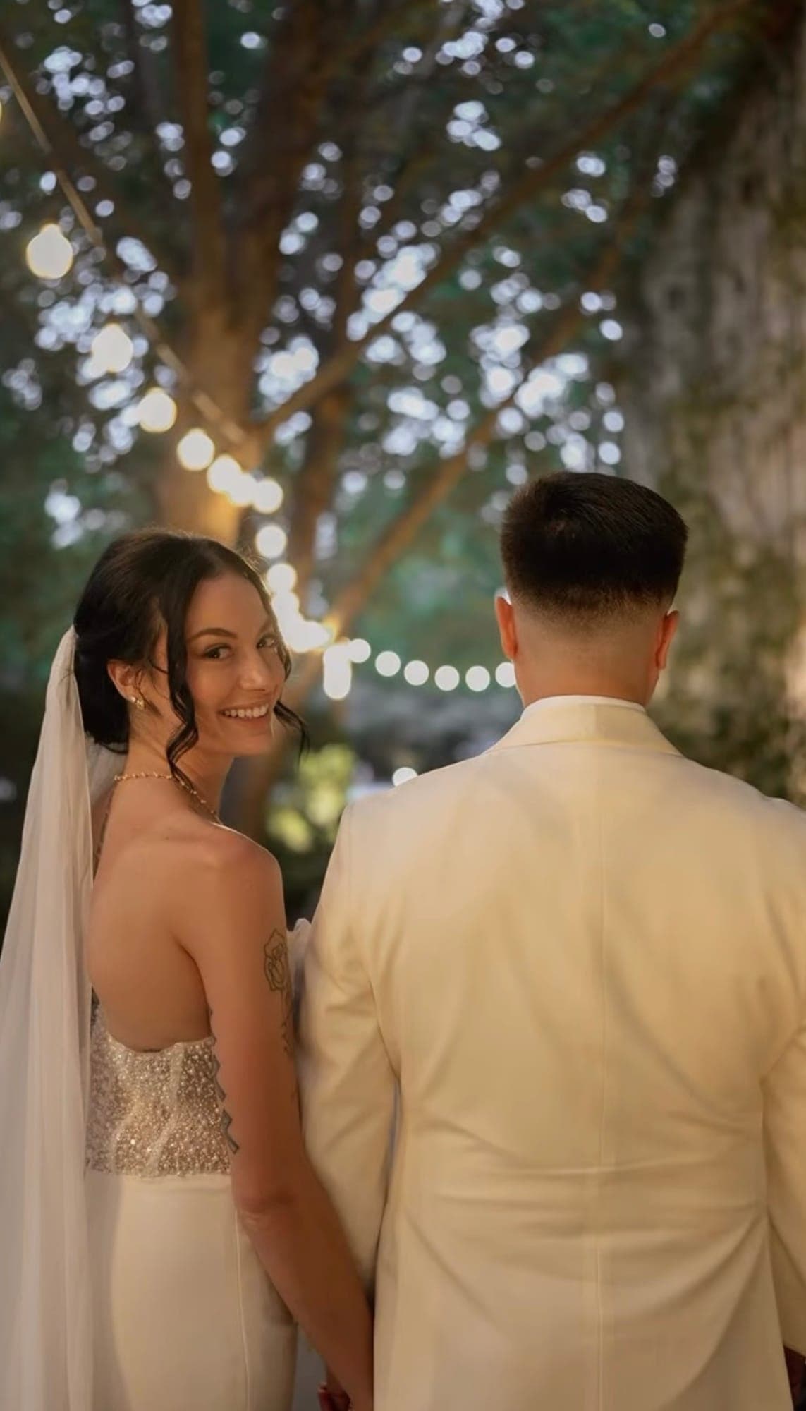 Novia mirando a cámara y sonriendo junto a su novio, entre las luces de la fiesta al atardecer